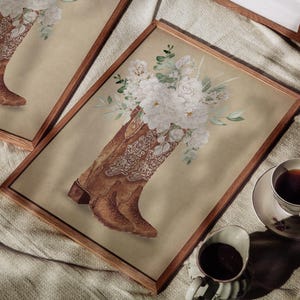 May include: A pair of brown cowboy boots with white floral embroidery, arranged in a vase with white flowers. The boots are displayed in a wooden frame on a beige fabric surface.