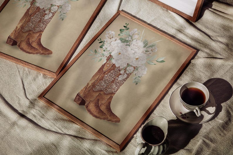 May include: A pair of brown cowboy boots with white floral embroidery, arranged in a vase with white flowers. The boots are displayed in a wooden frame on a beige fabric surface.