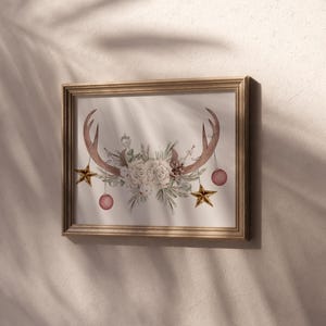 May include: A framed print of a watercolor illustration featuring a pair of antlers with a floral arrangement of white roses and greenery. The antlers are adorned with gold stars and pink ornaments.