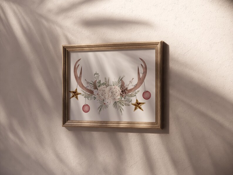 May include: A framed print of a watercolor illustration featuring a pair of antlers with a floral arrangement of white roses and greenery. The antlers are adorned with gold stars and pink ornaments.