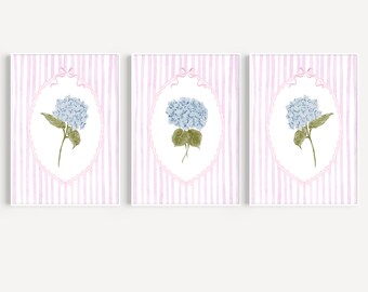 May include: Set of three framed art prints featuring watercolor illustrations of blue hydrangea flowers. Each print has a white frame, a pink and white striped background, and a decorative oval frame around the flower.