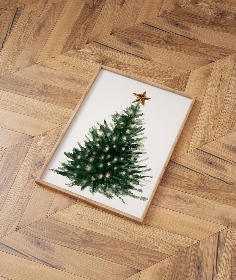 May include: A framed watercolor painting of a green Christmas tree with a gold star on top. The tree is surrounded by white space and the background is a light wood floor.