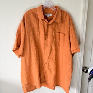 May include: A short-sleeved button-down shirt in a solid burnt orange color. The shirt features a standard collar, a single chest pocket on the left side, and a full-length front button closure. The fabric has a visible textured, linen-like weave.