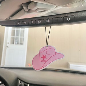 May include: Pink cowboy hat car air freshener hanging from the rearview mirror of a car. The hat has a red star on the front.