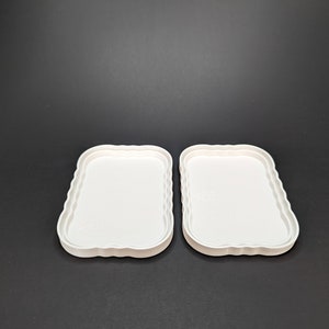 May include: Two white plastic trays with scalloped edges. The trays are empty and are sitting on a black surface.