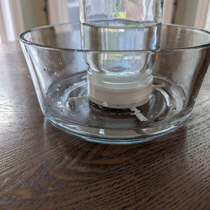 May include: A clear glass jar filled with water sits on a clear glass dish with a white plastic ring around the jar. The jar and dish are on a wooden surface.