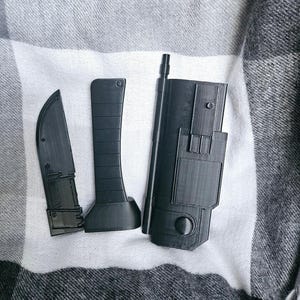 May include: Three black plastic utility knives with different blade shapes and sizes. The knives are on a gray and white plaid fabric background.