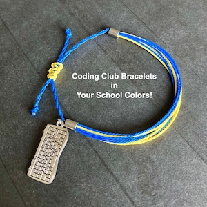 May include: A blue and yellow Coding Club bracelet with a keyboard charm. The bracelet features multiple strands of blue and yellow cord, with a silver-colored keyboard charm hanging from the adjustable knot. The text "Coding Club Bracelets in Your School Colors!" is visible.