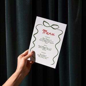 May include: A white menu card with red and green text and design elements is held by a person. The menu lists starters, main courses, and desserts. The menu includes Goat Cheese and Cranberry Crostini, Maple Glazed Ham, and Gingerbread Cheesecake Bars.
