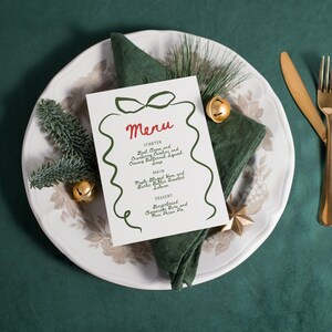 May include: A festive table setting featuring a white plate with a floral design, a green napkin, and a menu card. The menu card has the word "Menu" in red script and a green ribbon design. Gold cutlery and bells add a touch of elegance.