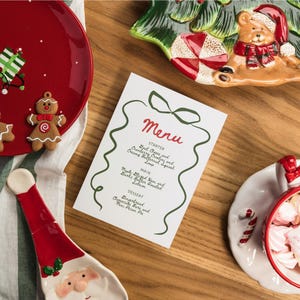 May include: A festive Christmas-themed arrangement featuring a menu card with "Menu" in red script. The card is surrounded by green decorative lines. A red plate with a gingerbread man and a Santa-shaped spoon are also visible.