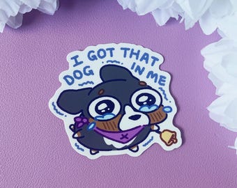 May include: A cartoon sticker of a dog with big eyes and a sad expression. The dog is wearing a purple bandana and a bell. The text "I got that dog in me" is written around the dog.