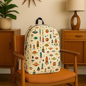 Puede incluir: Una mochila de color crema con un patrón de cohetes y planetas de estilo retro. La mochila está sentada en una silla de madera con un cojín naranja.