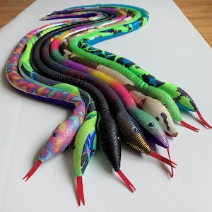 Pode incluir: Um conjunto de dez brinquedos de cobra de pelúcia coloridos e estampados. As cobras estão dispostas em uma linha curva, com as cabeças das cobras voltadas para o lado direito da imagem. As cobras são feitas de um material macio e de pelúcia e têm uma variedade de cores e padrões.