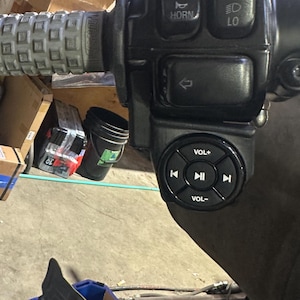 Puede incluir: Primer plano del manillar de una motocicleta con empuñadura texturizada y panel de control. El panel tiene botones para luces, bocina y controles de audio. Espejo retrovisor en el fondo.