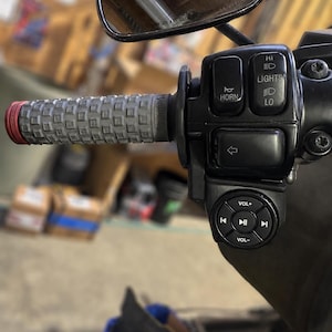 May include: Close-up of a motorcycle handlebar with a gray grip and red end cap. Black control panel with horn, lights, and turn signal buttons. A small round control panel with volume and media control buttons is also visible.