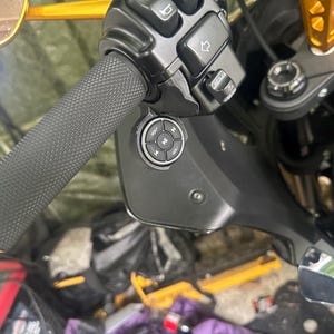 May include: Close-up of a motorcycle handlebar with a textured black grip and control buttons. The handlebar features a round control panel with volume and track selection buttons. The motorcycle has gold accents and a black fairing.