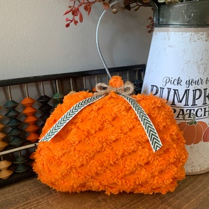 May include: An orange plush pumpkin decoration with a textured surface and a decorative ribbon. The pumpkin is adorned with a small stem and a bow. A metal container with fall foliage and a sign that says "Pick your own PUMPKIN PATCH" is in the background.
