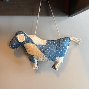 May include: A handmade, stuffed cow ornament with a patchwork design. The cow is primarily blue with white polka dots and cream-colored patches. It hangs from a jute string, showcasing a rustic, country aesthetic.
