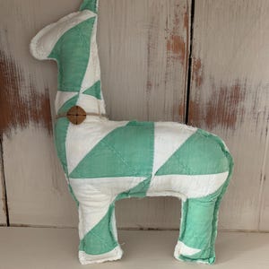 May include: A green and white patchwork fabric llama toy. The llama is standing on a white surface.
