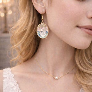 May include: Gold-toned jewelry set featuring a swan-themed earring and a delicate necklace. The oval-shaped earring showcases a swan design in pastel colors. The necklace has a small pearl pendant. The jewelry is worn by a person with blonde hair.