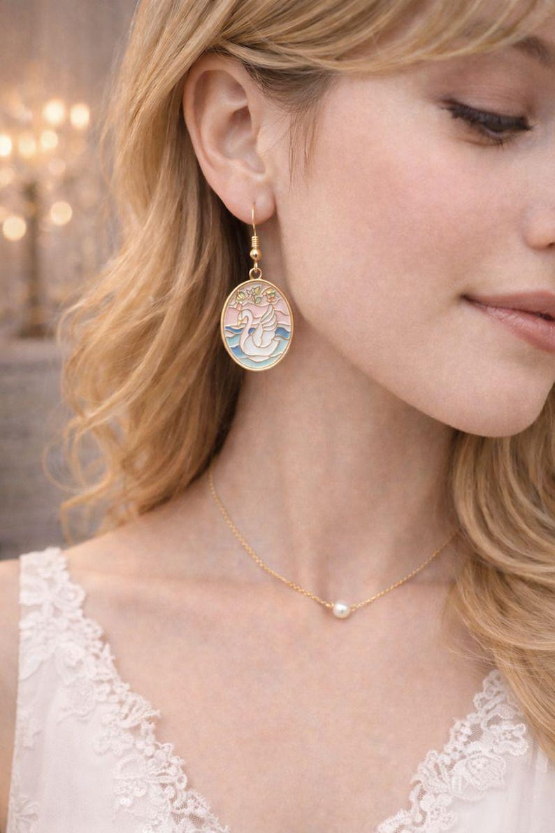 May include: Gold-toned jewelry set featuring a swan-themed earring and a delicate necklace. The oval-shaped earring showcases a swan design in pastel colors. The necklace has a small pearl pendant. The jewelry is worn by a person with blonde hair.