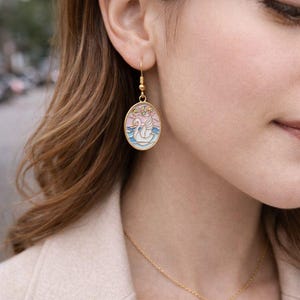 May include: Gold-toned oval earrings with a swan design. The earrings feature a gold hook and a pendant with a swan on a pink and blue background. A gold necklace with a single pearl is also visible.