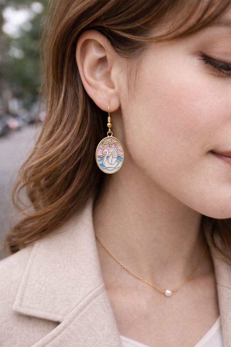 May include: Gold-toned oval earrings with a swan design. The earrings feature a gold hook and a pendant with a swan on a pink and blue background. A gold necklace with a single pearl is also visible.