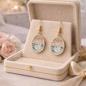 May include: Gold-tone earrings with an oval pendant featuring a white swan on blue water, set against a pink and light blue sky. The earrings are displayed in an open, cream-colored jewelry box with a gold trim.