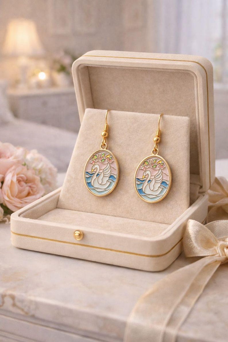 May include: Gold-tone earrings with an oval pendant featuring a white swan on blue water, set against a pink and light blue sky. The earrings are displayed in an open, cream-colored jewelry box with a gold trim.