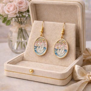 May include: Gold-toned dangle earrings featuring an oval design with a swan illustration. The earrings are set against a pink and blue background, with floral accents. Presented in a beige jewelry box, with a bouquet of pink roses in the background.