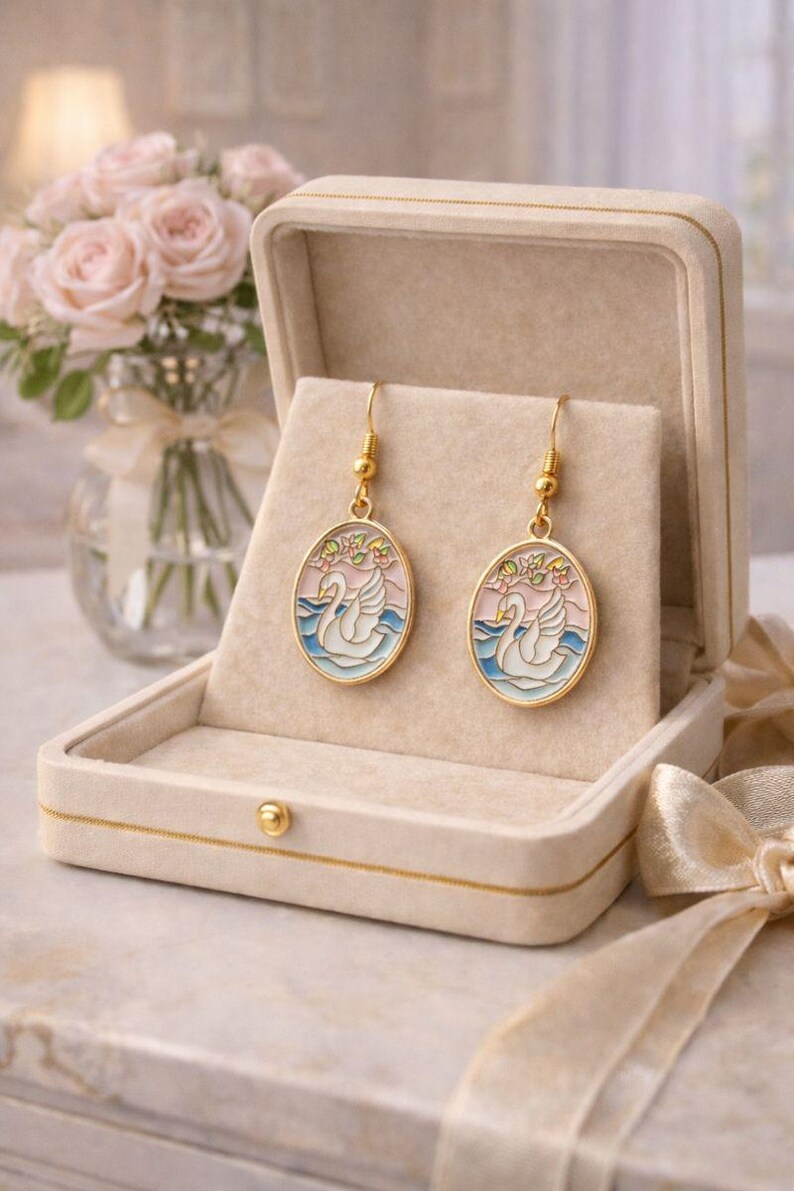 May include: Gold-toned dangle earrings featuring an oval design with a swan illustration. The earrings are set against a pink and blue background, with floral accents. Presented in a beige jewelry box, with a bouquet of pink roses in the background.