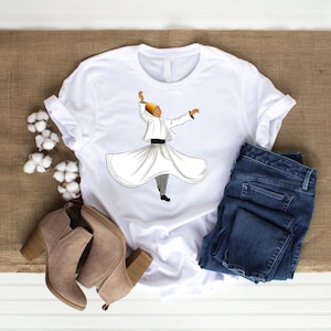 Può includere: Maglietta bianca con una grafica di una persona in una tunica bianca e un cappello giallo, che gira con le braccia distese. La maglietta è appoggiata su una superficie marrone con un paio di stivaletti marroni e un paio di jeans blu.