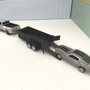 May include: A silver pickup truck towing a black dump trailer with a silver sports car on the ramp. The truck and trailer are both toy models.