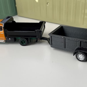 May include: A toy orange dump truck with a black bed and green wheels is towing a black utility trailer with a black bed and silver wheels.