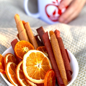 Peut inclure: Un bol blanc rempli de tranches d'orange s&eacute;ch&eacute;es et de rouleaux de cuir de fruits. Les rouleaux de cuir de fruits sont de diff&eacute;rentes nuances de brun et de rouge. Les tranches d'orange sont d'une couleur orange vif. Une tasse blanche avec un motif de pomme rouge est en arri&egrave;re-plan.