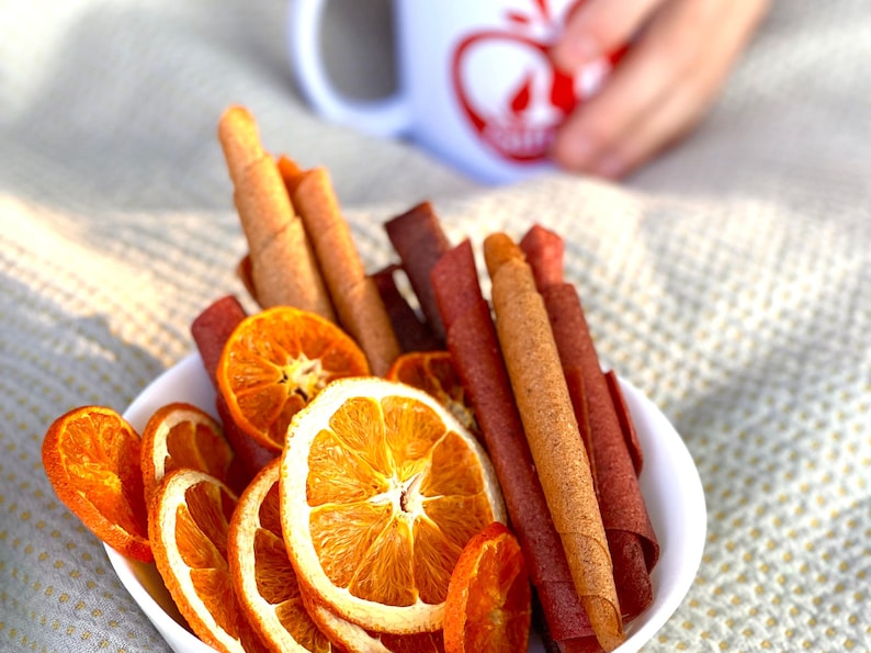 Peut inclure: Un bol blanc rempli de tranches d'orange s&eacute;ch&eacute;es et de rouleaux de cuir de fruits. Les rouleaux de cuir de fruits sont de diff&eacute;rentes nuances de brun et de rouge. Les tranches d'orange sont d'une couleur orange vif. Une tasse blanche avec un motif de pomme rouge est en arri&egrave;re-plan.