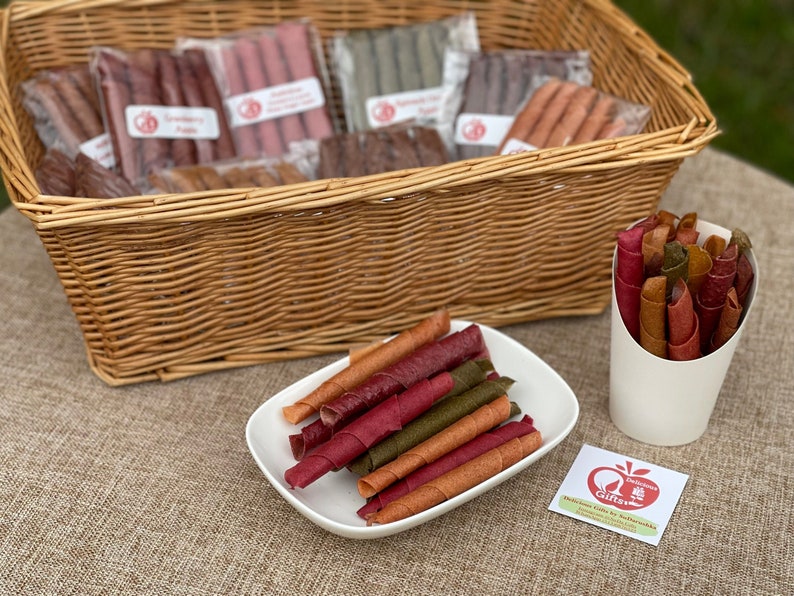 Peut inclure: Un panier en osier rempli de rouleaux de fruits secs emball&eacute;s individuellement. Une assiette blanche et un gobelet en papier contiennent d'autres rouleaux de diff&eacute;rentes couleurs. Une petite &eacute;tiquette avec du texte et un logo de pomme est visible.