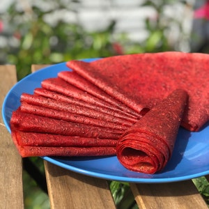 Peut inclure: Une assiette bleue pr&eacute;sente plusieurs feuilles de cuir de fruits rouge, &eacute;tal&eacute;es et enroul&eacute;es. Le cuir de fruits a un aspect textur&eacute; et une couleur rouge fonc&eacute;. L'assiette est pos&eacute;e sur une surface en bois avec un arri&egrave;re-plan vert flou.