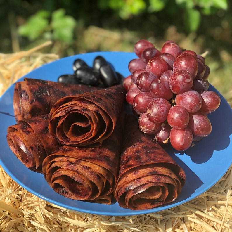 Peut inclure: Une assiette bleue pr&eacute;sente des rouleaux de cuir de fruits, des raisins noirs et des raisins rouges. Le cuir de fruits est brun fonc&eacute;, roul&eacute; en spirales. Les raisins sont d'un rouge profond et noir, cr&eacute;ant une pr&eacute;sentation color&eacute;e et app&eacute;tissante.