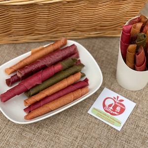 Peut inclure: Assortiment de rouleaux de cuir de fruits color&eacute;s, aux teintes rouge, orange et verte, pr&eacute;sent&eacute;s sur une assiette blanche et dans un c&ocirc;ne en papier blanc. Une petite &eacute;tiquette avec du texte et un logo est &eacute;galement visible.