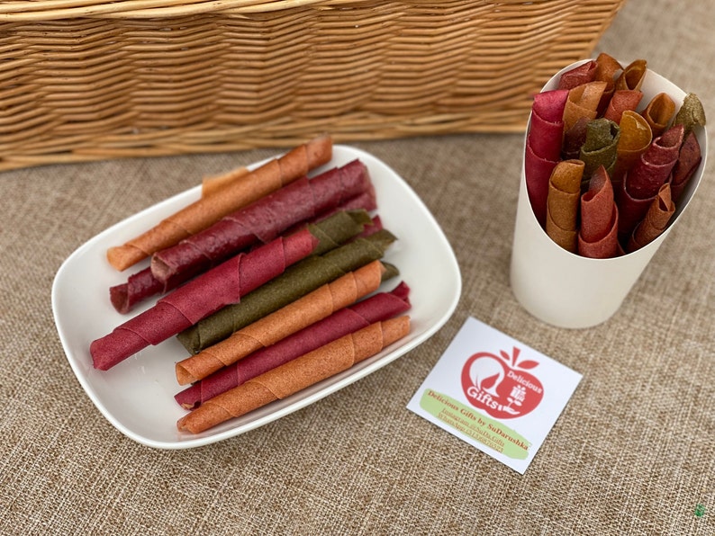 Peut inclure: Assortiment de rouleaux de cuir de fruits color&eacute;s, aux teintes rouge, orange et verte, pr&eacute;sent&eacute;s sur une assiette blanche et dans un c&ocirc;ne en papier blanc. Une petite &eacute;tiquette avec du texte et un logo est &eacute;galement visible.