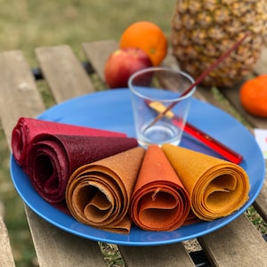 Peut inclure: Une assiette bleue pr&eacute;sente des rouleaux de cuir de fruits aux teintes rouges, marron et jaunes. Une orange, une pomme et un ananas sont en arri&egrave;re-plan. Un verre et un agitateur rouge sont &eacute;galement sur l'assiette.