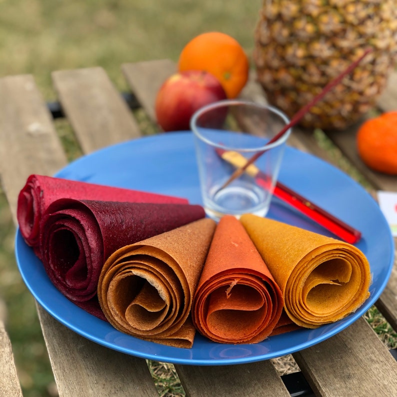 Peut inclure: Une assiette bleue pr&eacute;sente des rouleaux de cuir de fruits aux teintes rouges, marron et jaunes. Une orange, une pomme et un ananas sont en arri&egrave;re-plan. Un verre et un agitateur rouge sont &eacute;galement sur l'assiette.