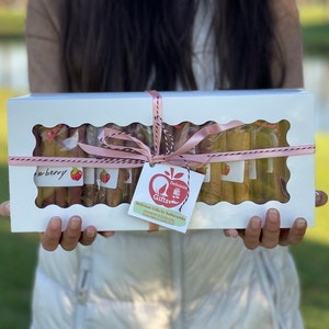 Peut inclure: Une bo&icirc;te cadeau rectangulaire blanche avec une fen&ecirc;tre transparente, attach&eacute;e avec de la ficelle rose et blanche. La bo&icirc;te contient plusieurs rouleaux emball&eacute;s, chacun &eacute;tiquet&eacute; avec un motif de fraise. Une &eacute;tiquette avec un logo de pomme rouge et du texte est suspendue au ruban.