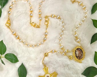 Puede incluir: Un rosario de perlas blancas con una cruz dorada y un colgante ovalado dorado con una imagen religiosa. El rosario está sobre una superficie blanca y peluda.