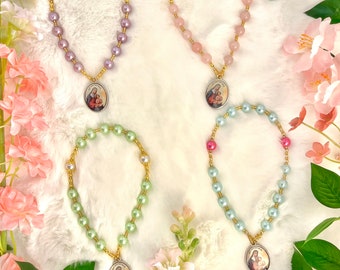 May include: Four different colored beaded necklaces with a gold charm featuring a religious image of Mary and Jesus. The necklaces are on a white fuzzy background with pink flowers.