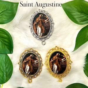May include: Three oval-shaped pendants with ornate gold and silver frames. Each pendant features a painting of Saint Augustine, a Catholic saint, holding a book and quill. The pendants are on a white fluffy background.