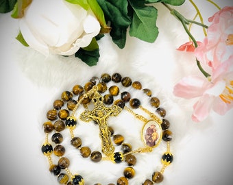 May include: A rosary with tiger eye beads, gold-colored metal accents, and a crucifix. The rosary features a small oval image and is displayed on a white, fluffy surface with floral accents.