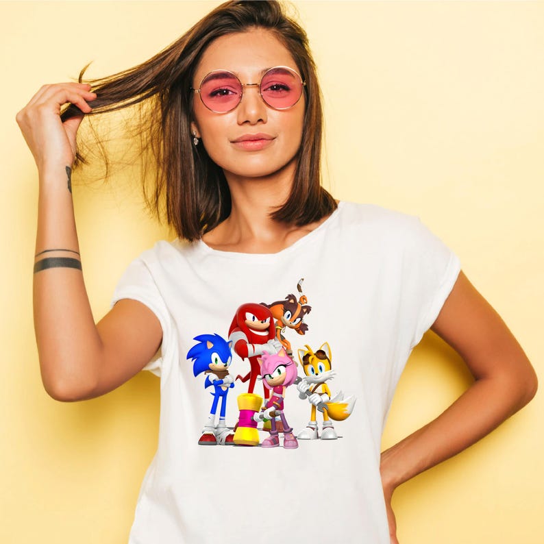 May include: White t-shirt featuring a colorful graphic of Sonic the Hedgehog and friends. The design includes Sonic, Knuckles, Amy Rose, Tails, and other characters. The shirt is made of a soft material.